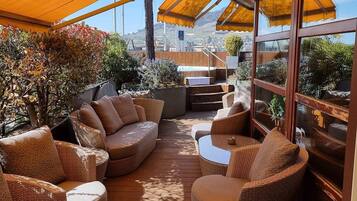 Terraza exterior con mobiliario de mimbre y suelo de madera, rodeada de toldos amarillos y plantas.