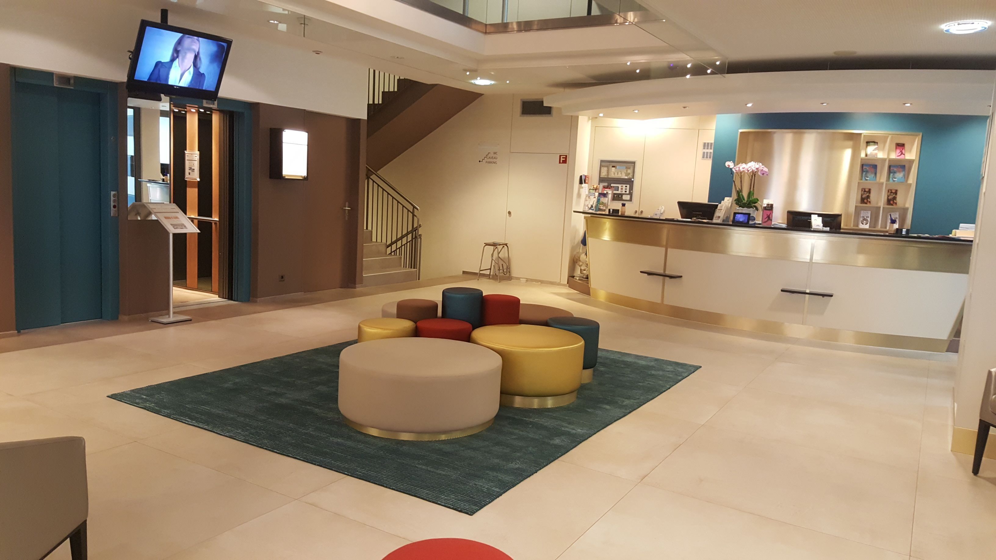 A modern lobby with a reception desk, a staircase, and a seating area with colorful ottomans.
