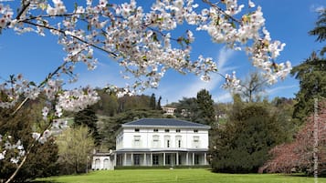 A large, white building with a symmetrical facade and a central entrance, surrounded by lush greenery and flowering trees.