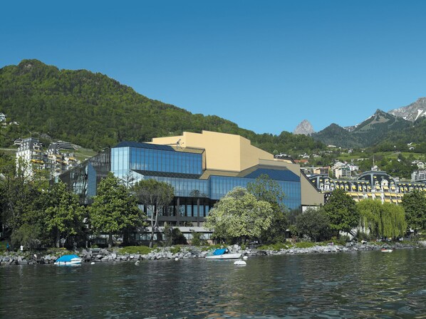 Un edificio moderno con fachada de vidrio, ubicado junto a un cuerpo de agua con barcos, rodeado de vegetación y montañas.