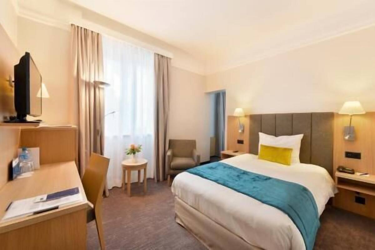 Ein Hotelzimmer mit Bett, Schreibtisch, Sessel, Fernseher und einem kleinen Tisch mit Lampe.