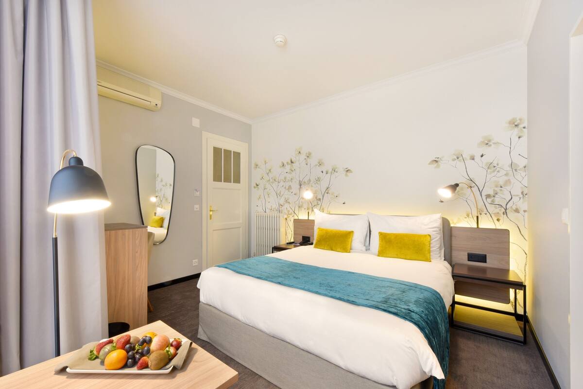 Ein Hotelzimmer mit einem ordentlich bezogenen Bett, Nachttischen, einer Obstplatte, einem Spiegel und einer Wand mit floralem Dekor.