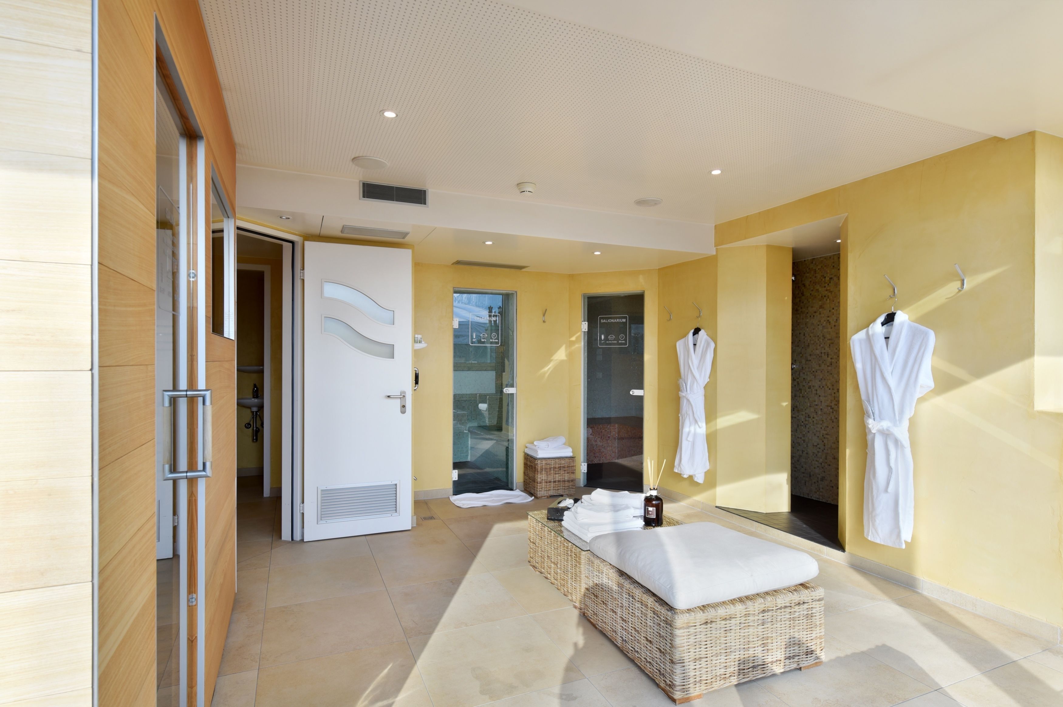 Une salle de spa moderne, dotée d'un grand canapé en osier, d'une douche en verre et de deux peignoirs blancs suspendus à des crochets.
