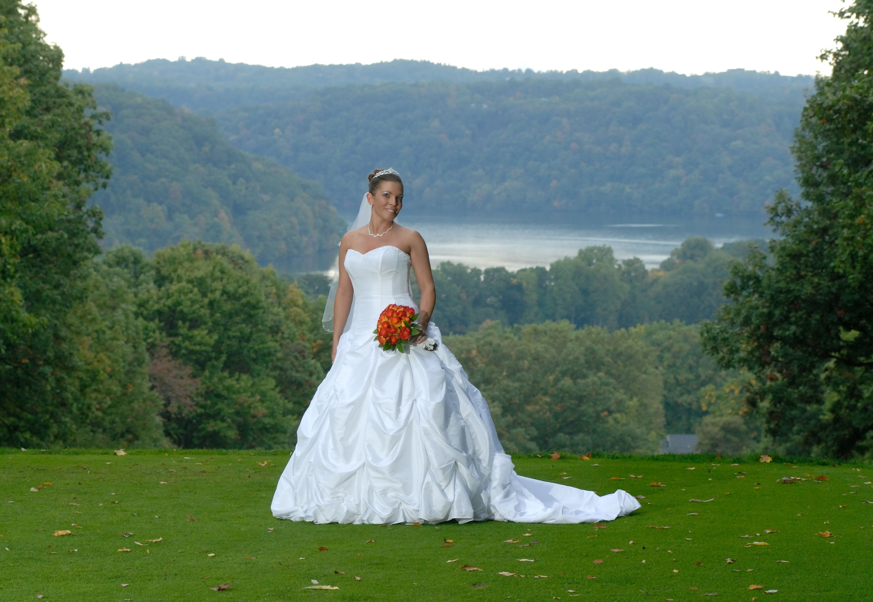 Una sposa in abito da cerimonia bianco con un bouquet, in piedi su un prato con uno specchio d'acqua e colline boscose sullo sfondo.