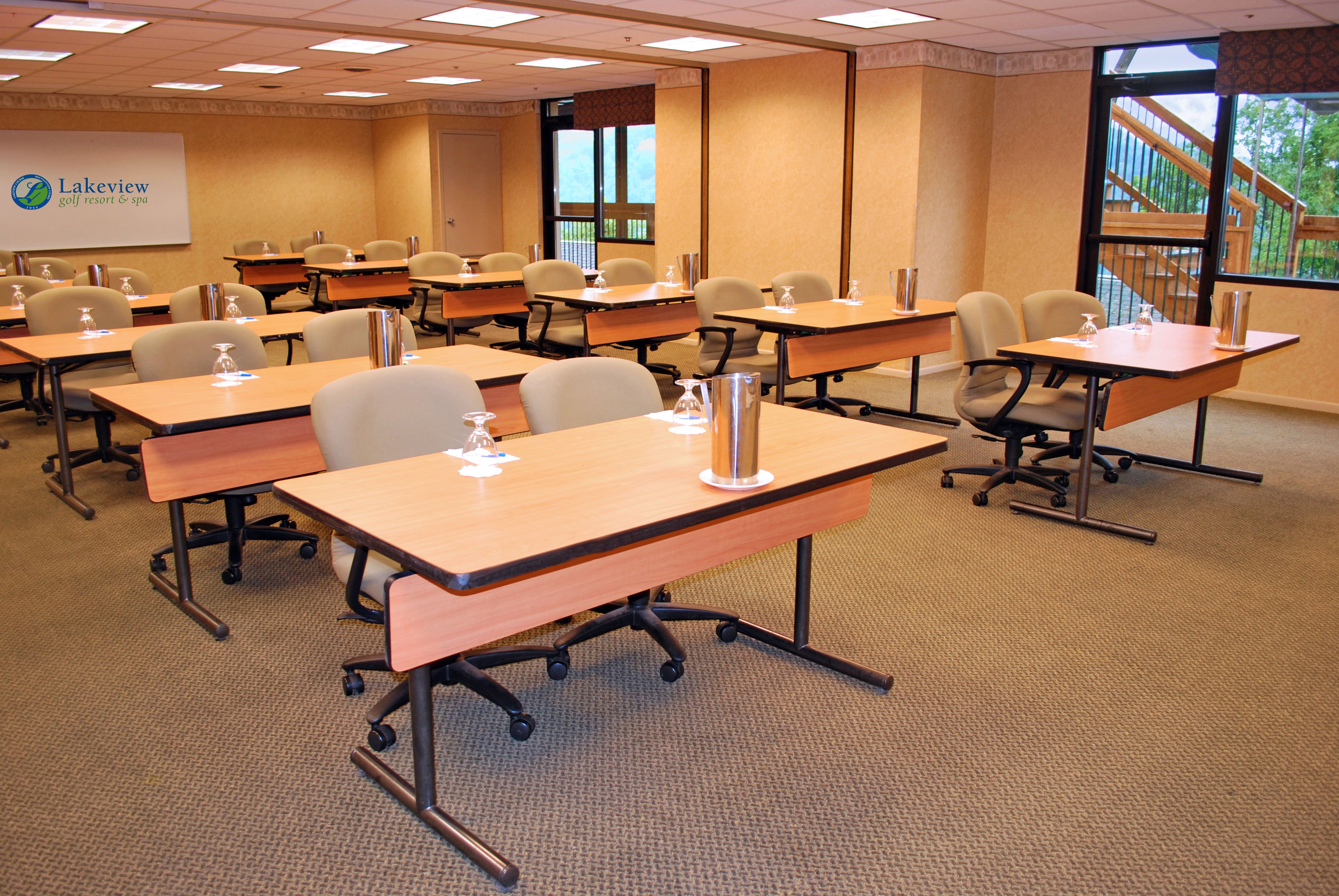 A conference room with multiple tables and chairs, a projector screen displaying 'Lakeview Golf Resort & Spa', and large windows with a view of outdoor stairs.