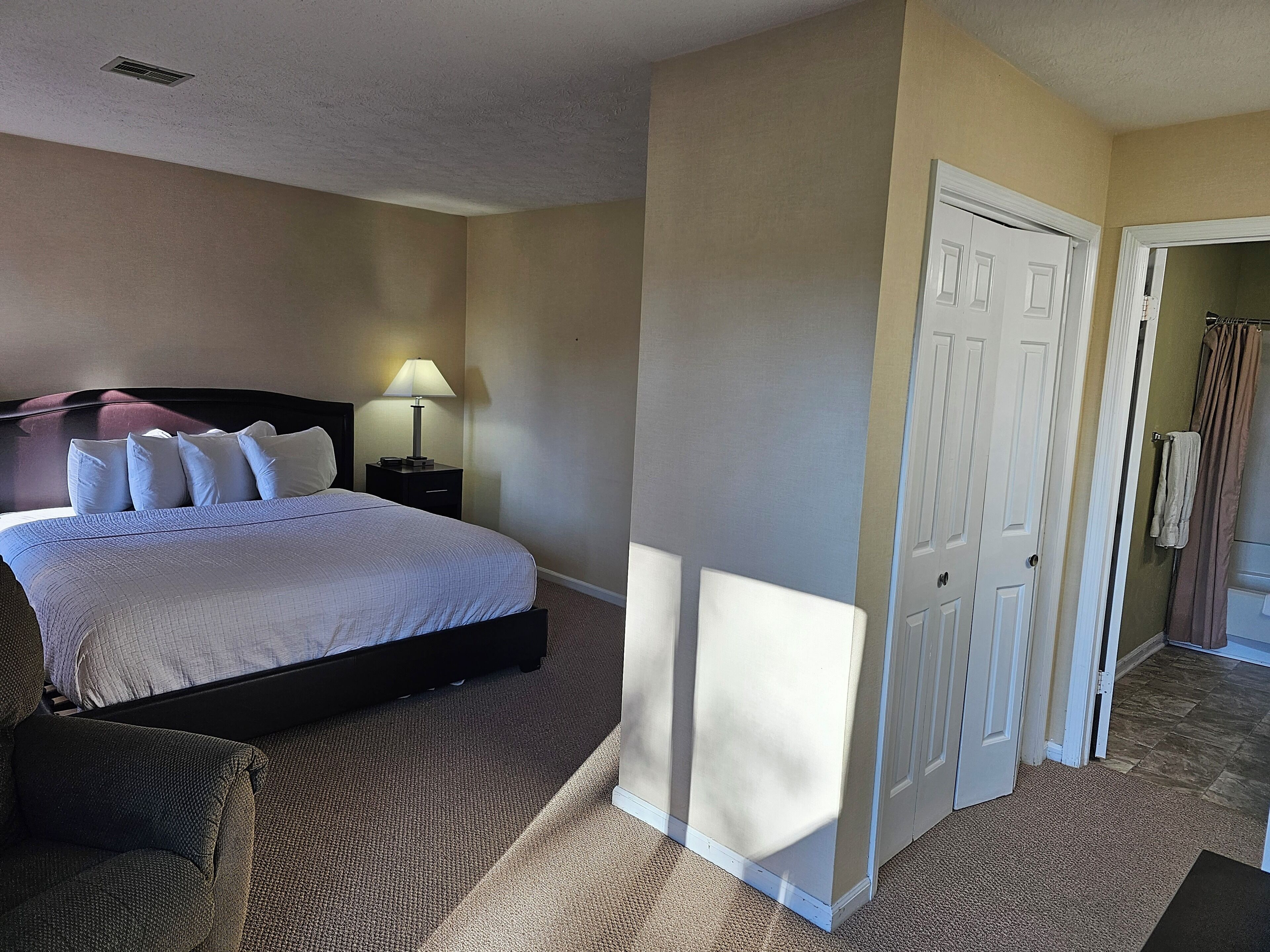 Habitación de hotel con cama, mesita de noche, lámpara y una puerta que da a otra habitación.