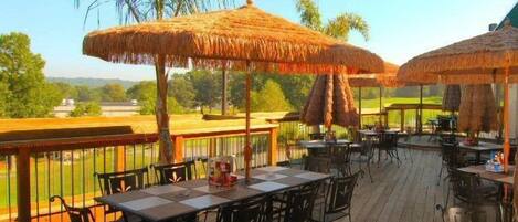 An outdoor deck with thatched umbrellas, wooden tables, and chairs, overlooking a green landscape.