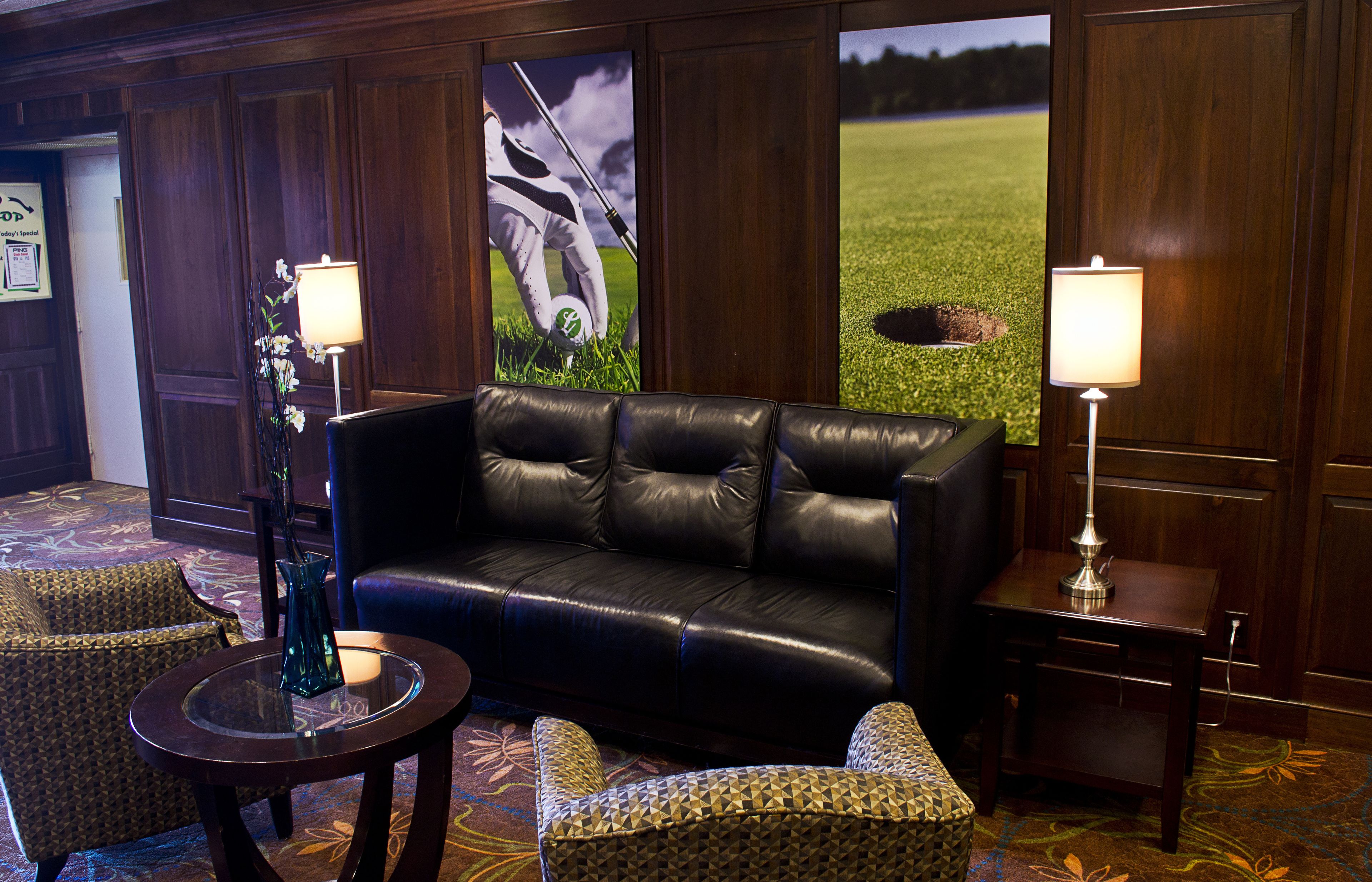 A lobby with a black leather sofa, two patterned armchairs, a glass table with a vase, and two wall-mounted lamps.