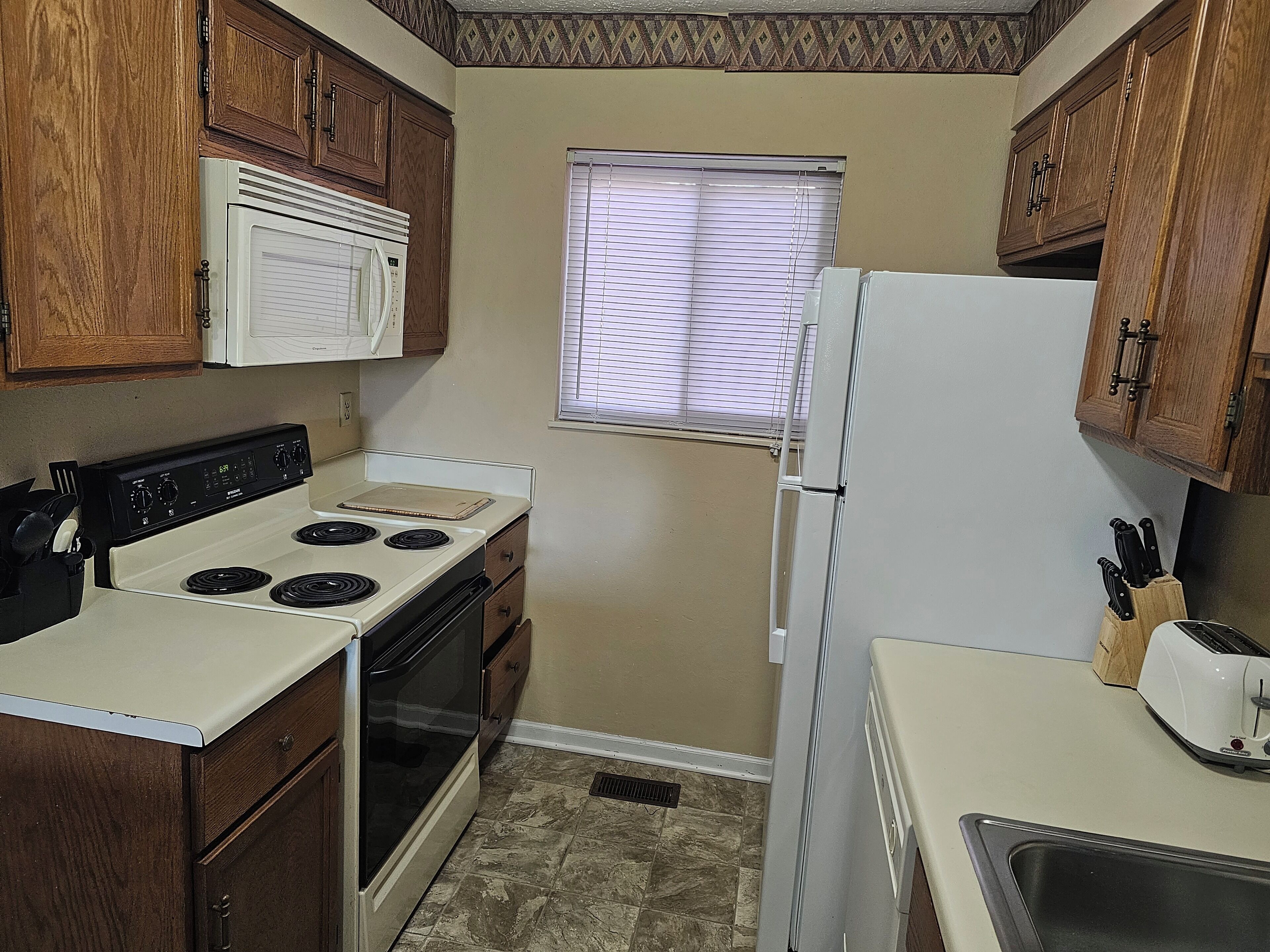 A compact kitchen with wooden cabinets, a white stove, a microwave, a refrigerator, and a window with blinds.
