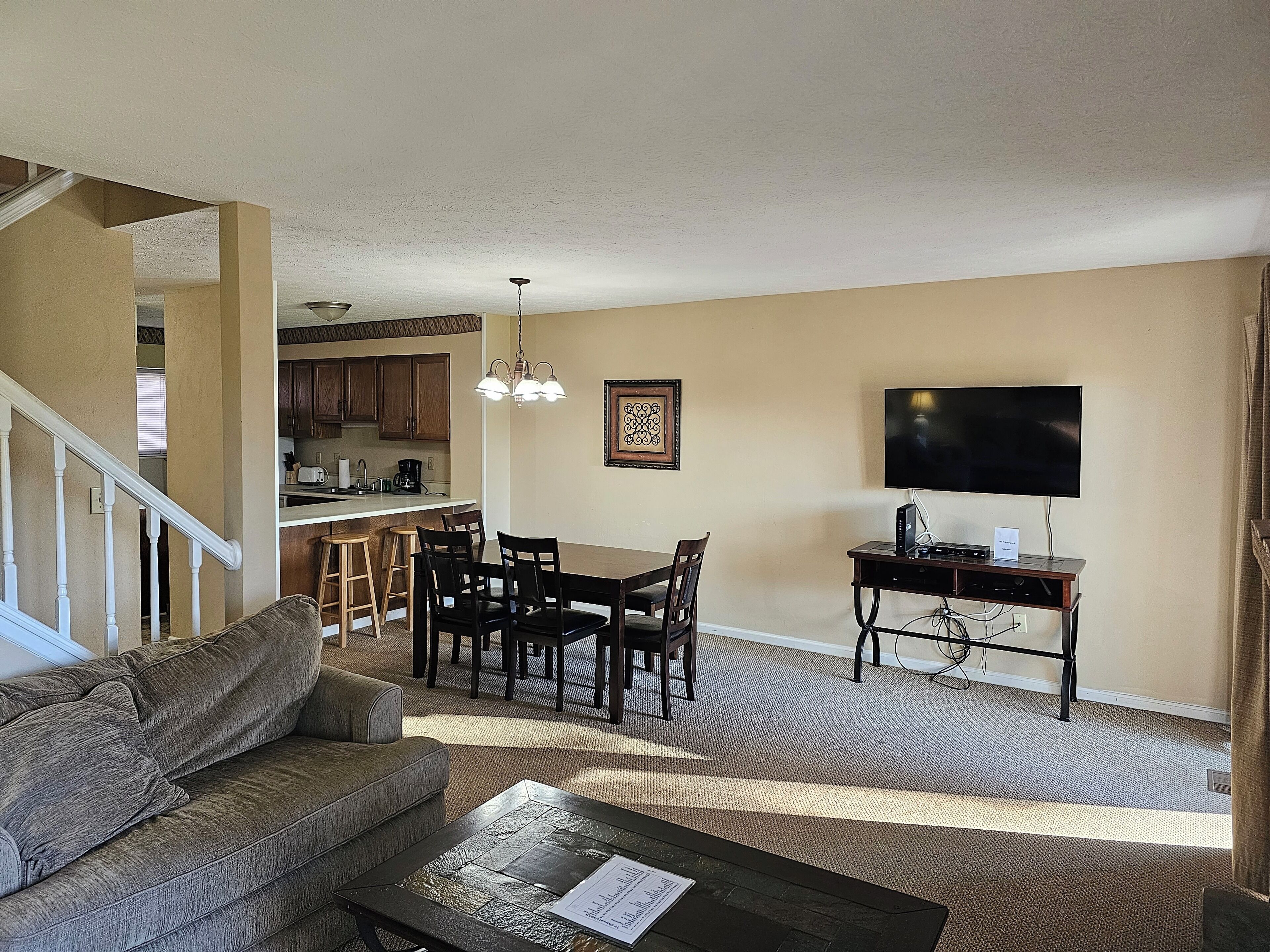 A living room with a sofa, dining table, chairs, a TV, and a kitchen in the background.
