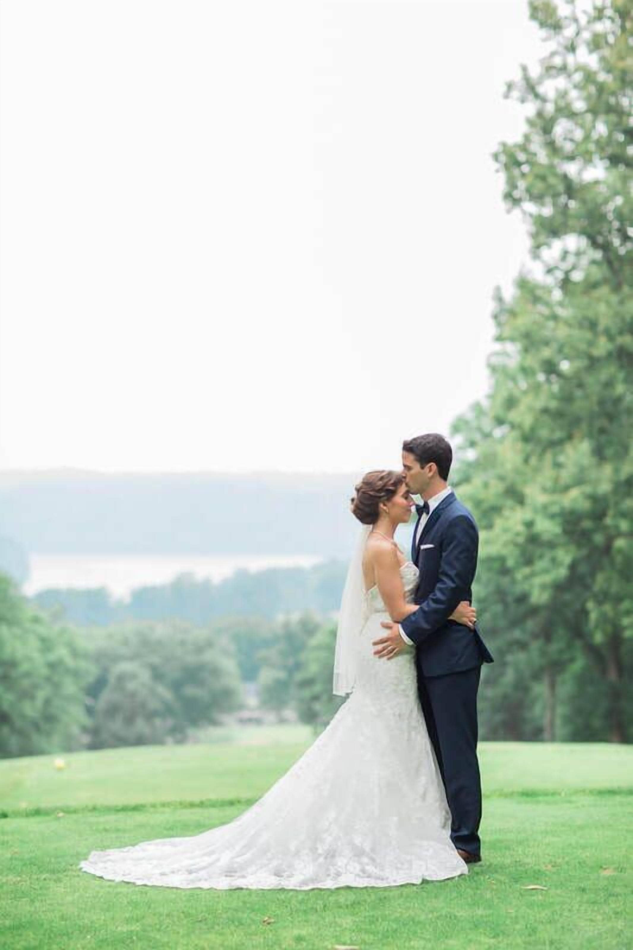 A couple embracing on a grassy field with a view of trees and a body of water in the background.