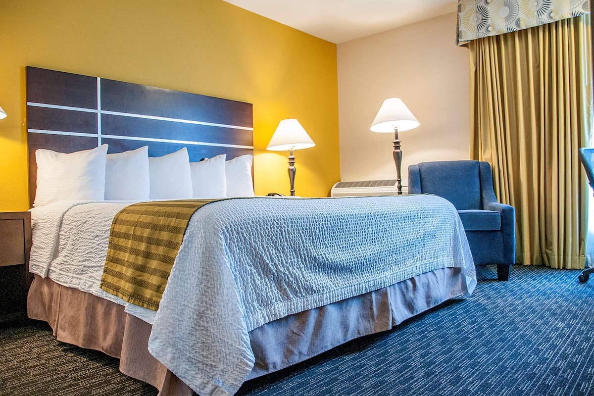 A hotel room with a bed, a chair, two lamps, and a large window with curtains.