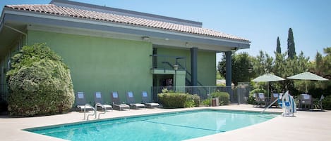 Uma piscina com espreguiçadeiras e guarda-sóis, um edifício com telhado em azulejos e um céu claro.