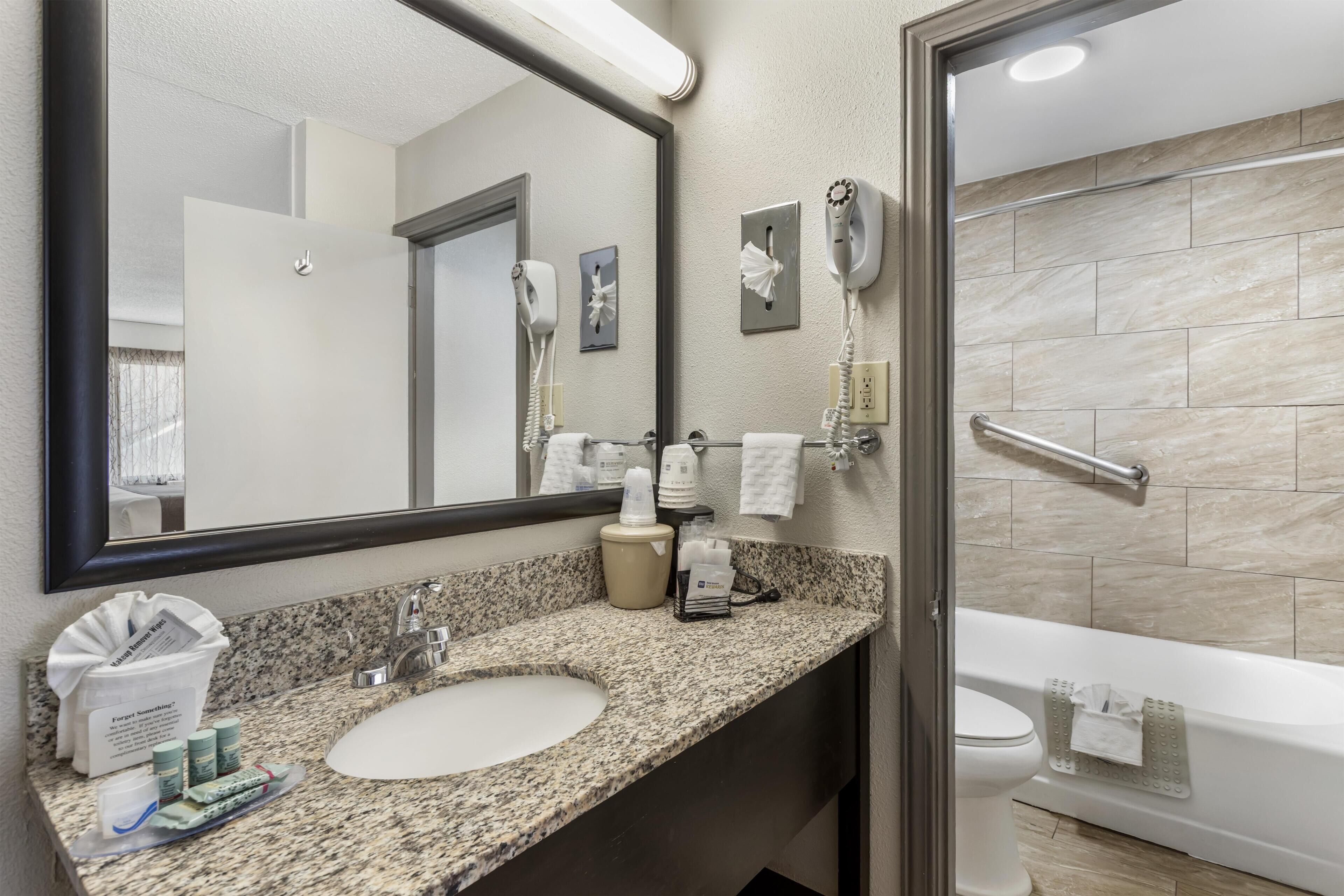 A bathroom with a granite countertop, a sink, a mirror, a hairdryer, a towel rack, and a bathtub.