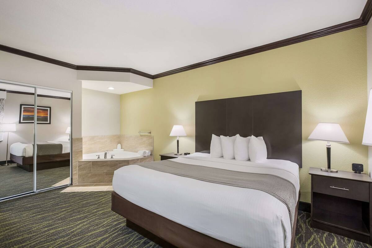 A hotel room with a large bed, a separate bathroom, a mirror, and a nightstand with a lamp.