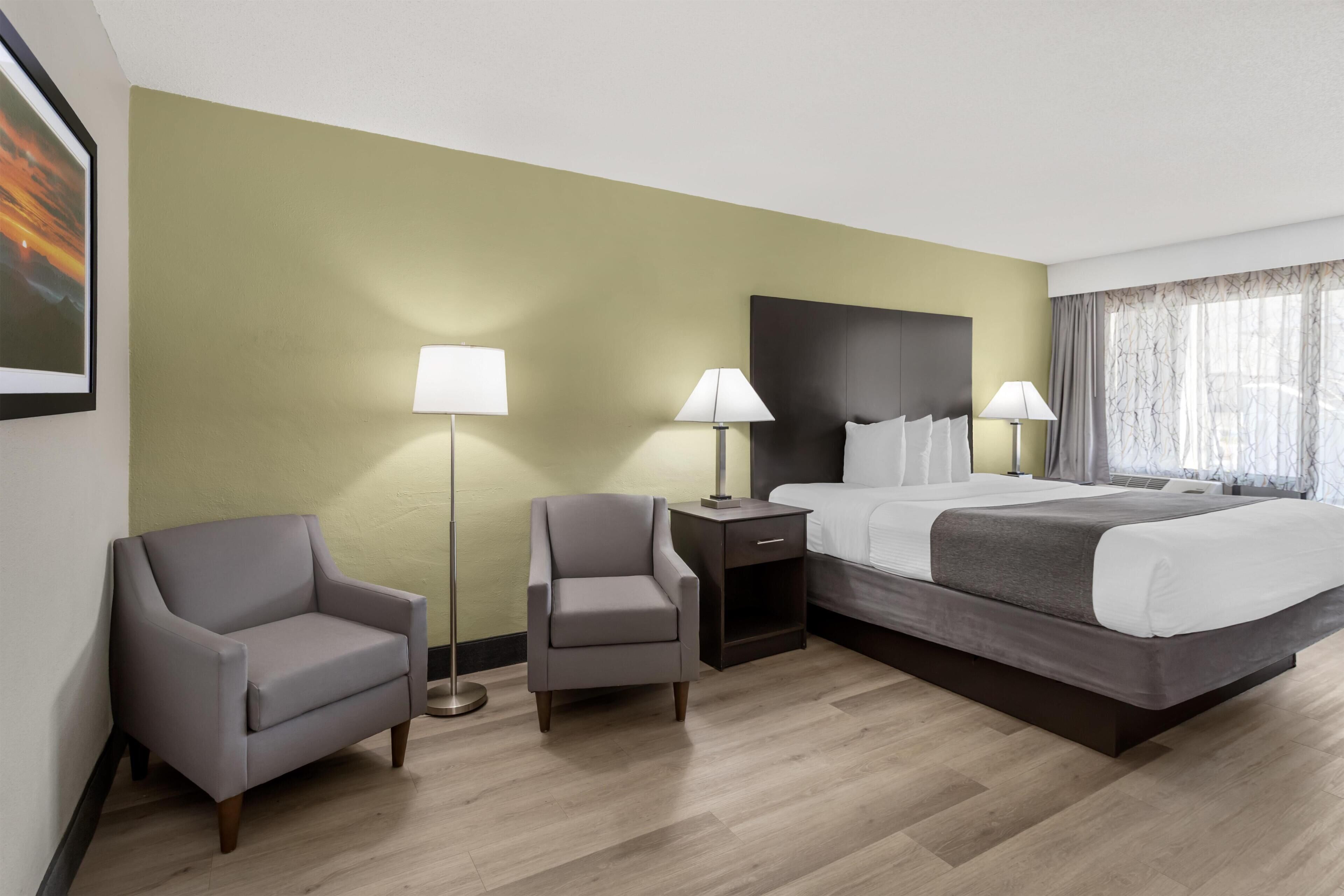 Hotellihuone, jossa on sänky, kaksi nojatuolia, yöpöytä ja suuri ikkuna, jossa on verhot.