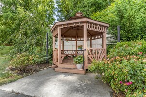 A wooden gazebo with a bench and potted plants, surrounded by lush greenery and flowers.