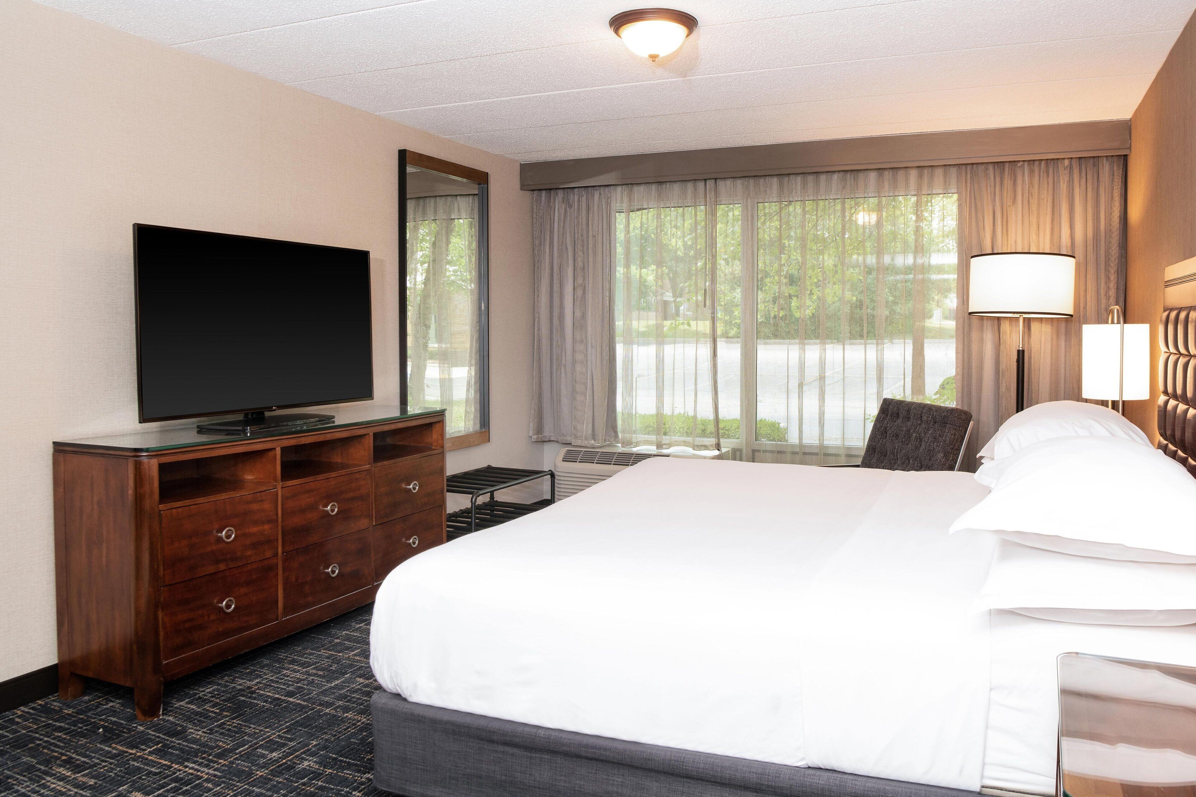 A hotel room with a large bed, a flat-screen TV on a wooden dresser, a desk with a chair, and a window with curtains.