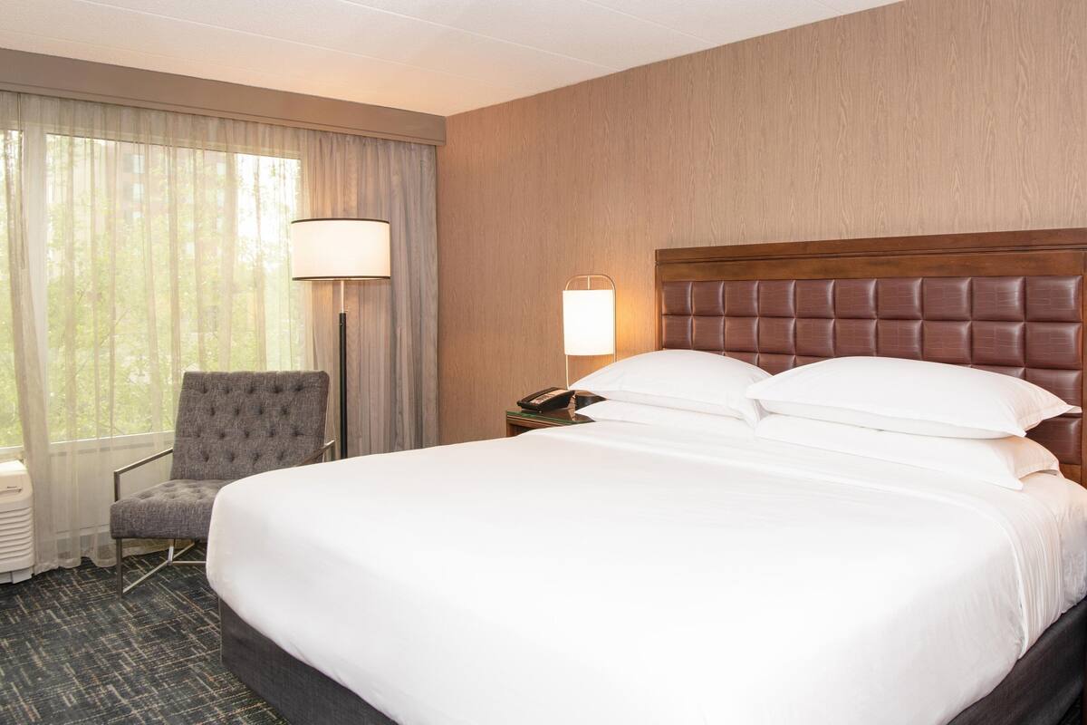 Habitación de hotel con una cama grande, una silla, una mesita de noche con lámpara y una ventana con cortinas.