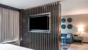 A hotel room with a large bed, a flat-screen TV mounted on a wooden panel, a small table, a nightstand with a lamp, and a blue rug. - Sheraton Ann Arbor Hotel (Ann Arbor)
