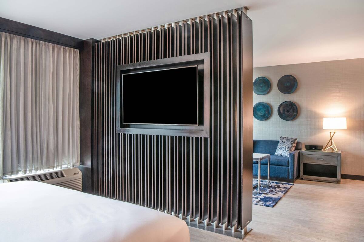 Habitación de hotel con una cama grande, un televisor de pantalla plana montado en un panel de madera, una mesita, una mesita de noche con lámpara y una alfombra azul.