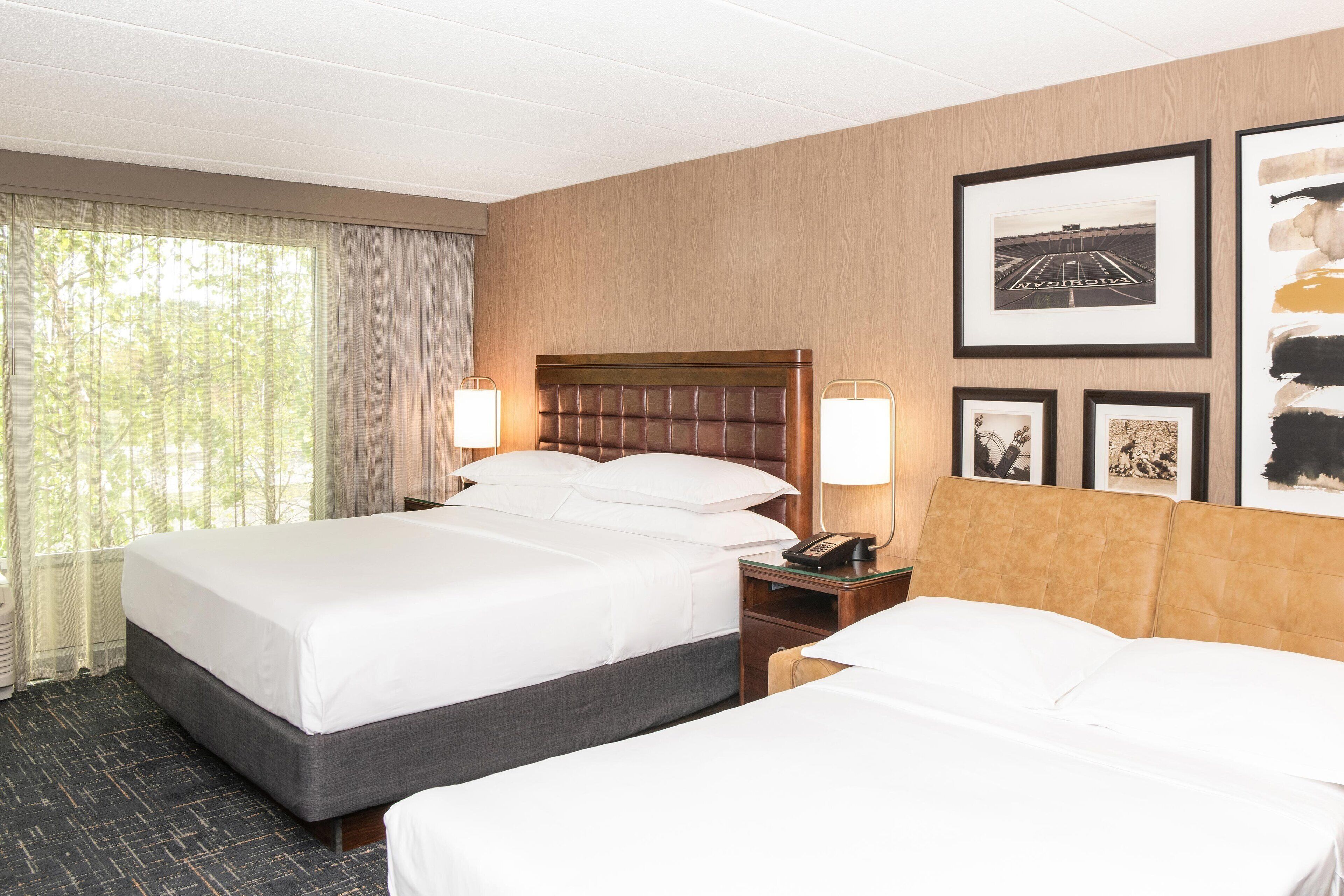 A hotel room with two beds, a wooden headboard, a nightstand with a lamp, and framed pictures on the wall.