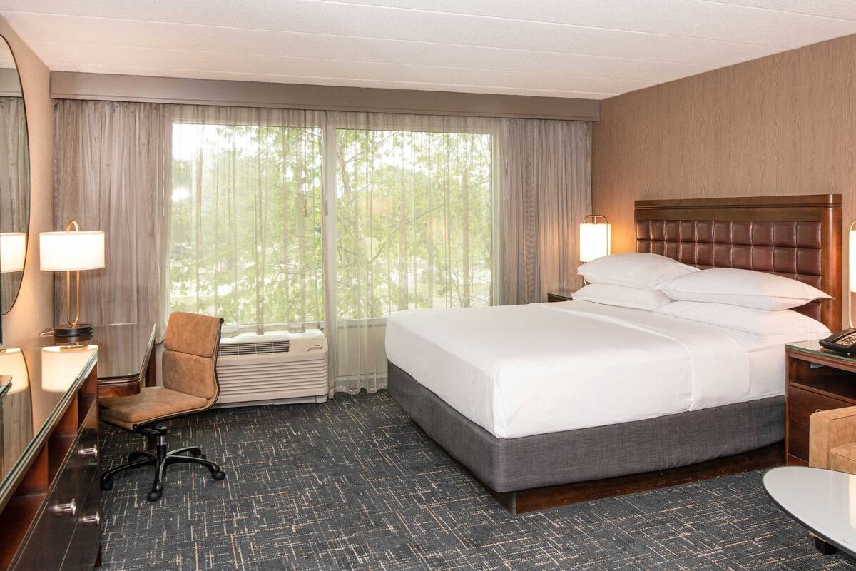 Habitación de hotel con una cama grande, una silla, un escritorio con lámpara y un ventanal con cortinas.