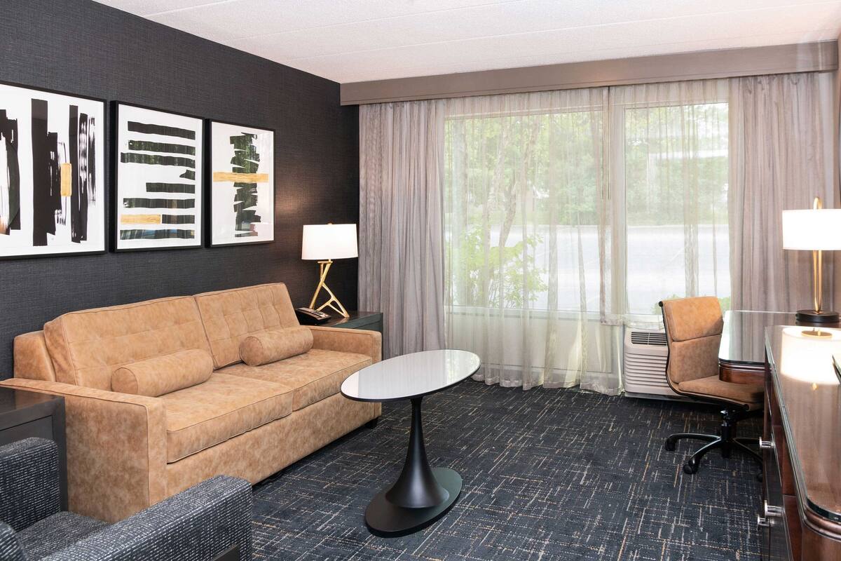 Una habitación de hotel con un sofá color beige, una mesa redonda, un escritorio con lámpara, una silla y obras de arte en la pared.
