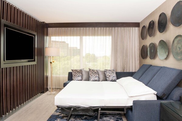 A modern hotel room with a bed, a sofa, a TV, and a city view.