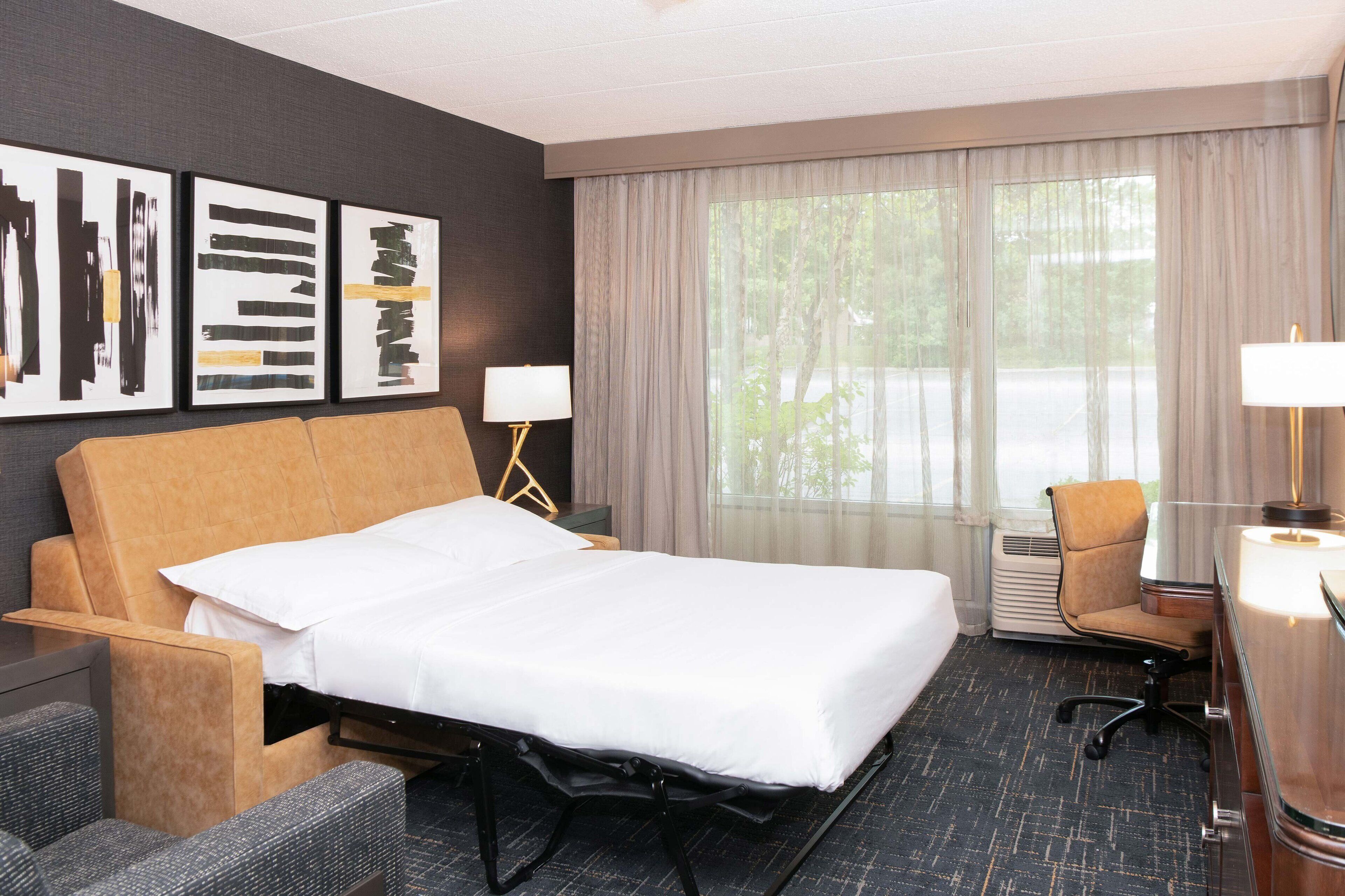 A hotel room with a bed, a desk, a chair, a lamp, and artwork on the wall.
