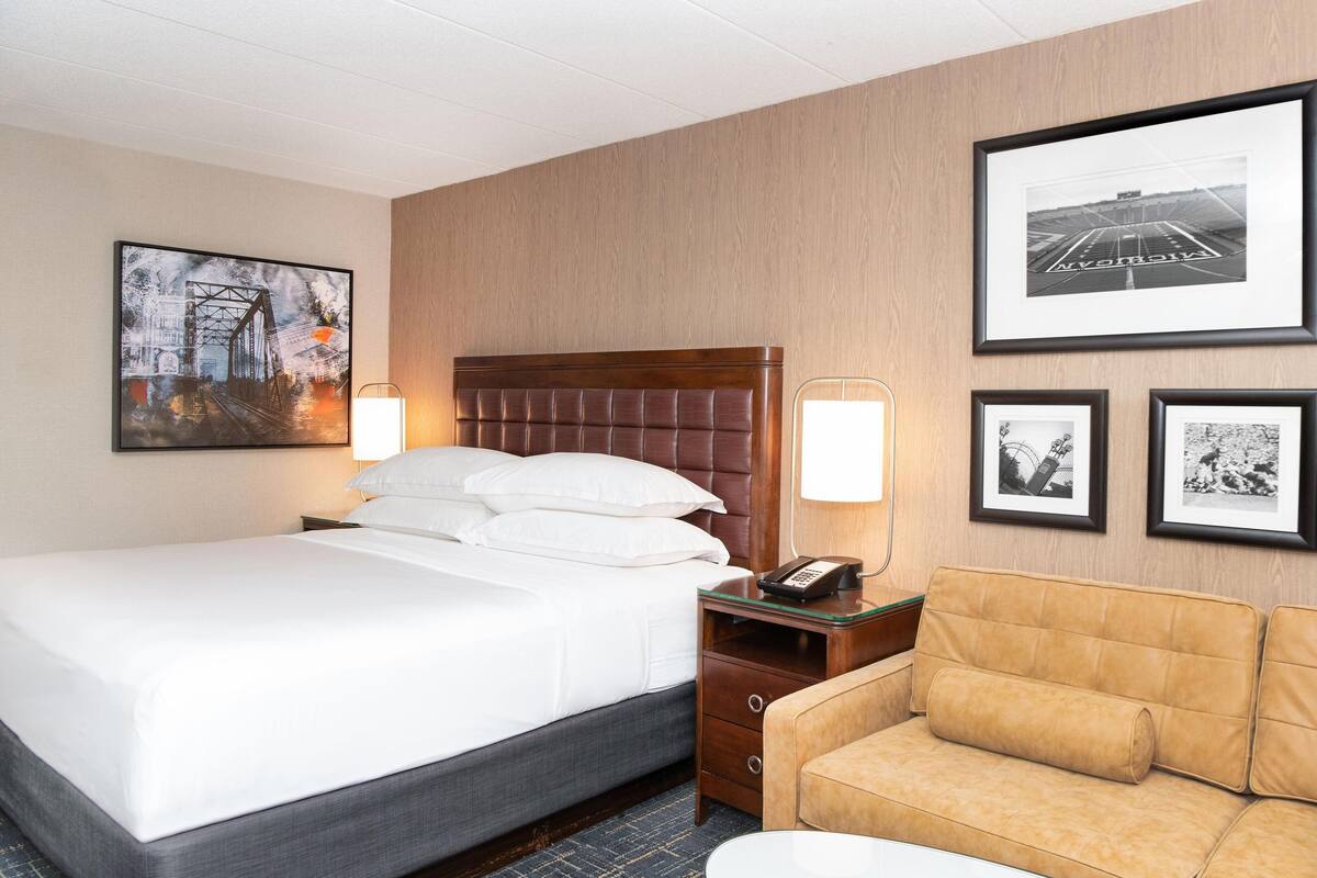 Una habitación de hotel con una cama grande, cabecera de madera, mesita de noche con lámpara, un sofá y cuadros en la pared.