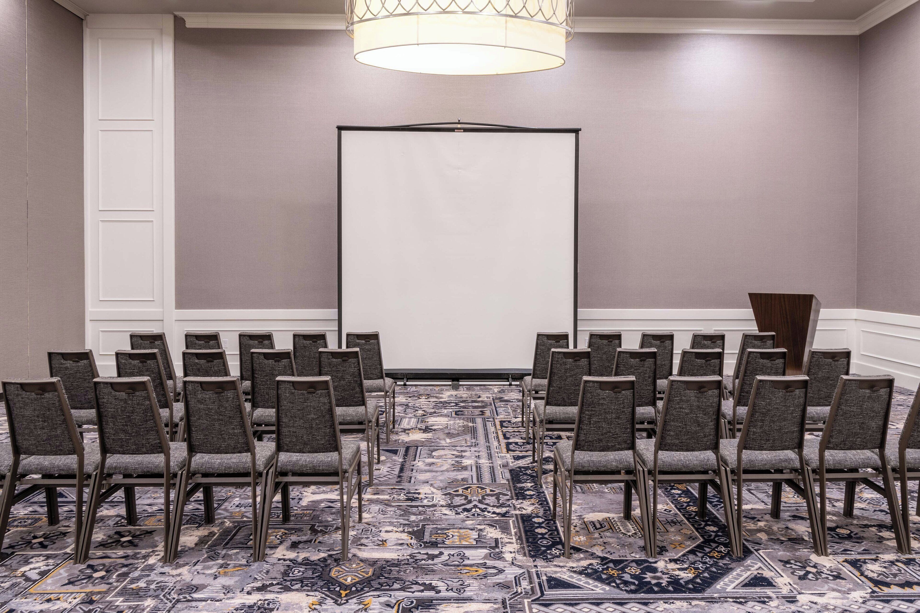 Une salle de conférence avec des rangées de chaises disposées face à un écran de projection vide, un tapis à motifs et un pupitre en bois.