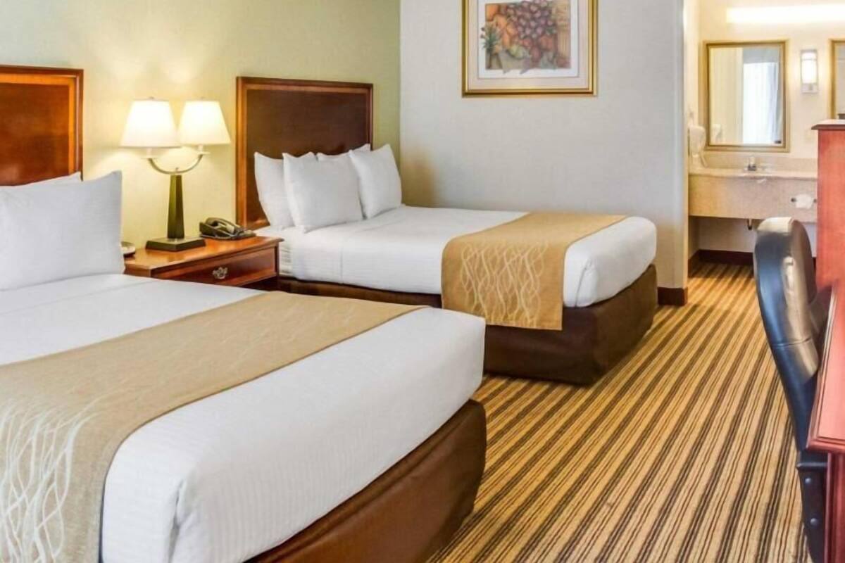 A hotel room with two beds, a desk, a chair, a lamp, a framed picture, and a bathroom mirror.