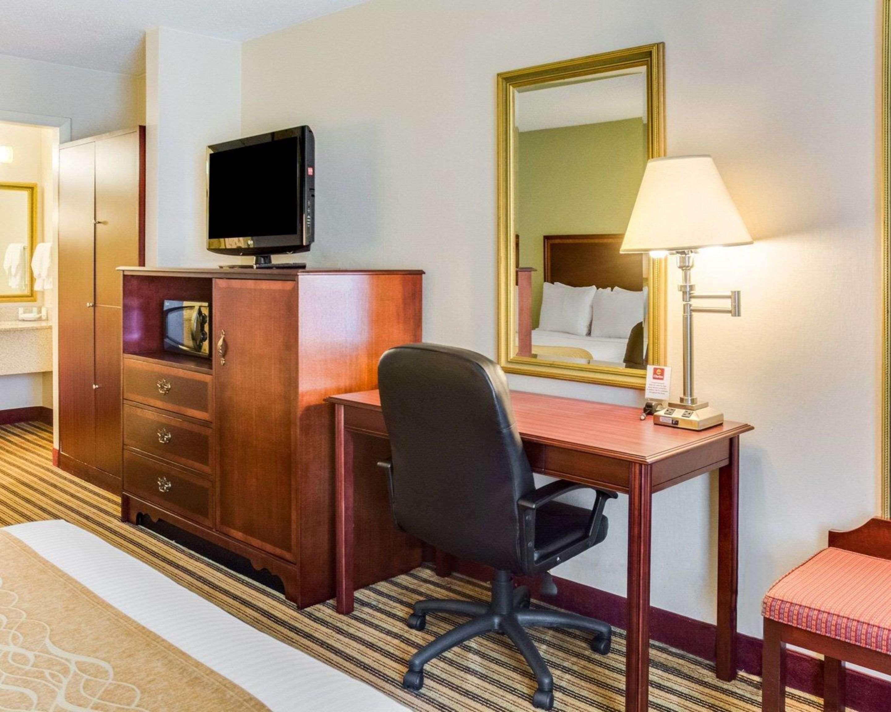 A hotel room with a wooden desk, a black office chair, a television, a bedside lamp, and a large mirror.