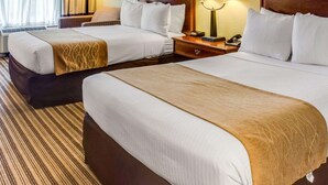 A hotel room with two beds, each with a wooden headboard, white bedding, and a brown throw blanket. There are bedside lamps, a framed picture on the wall, and a window with curtains. - Clarion Inn Fredericksburg North (Fredericksburg)