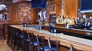 A bar area with a long counter, bar stools, a television mounted on the wall, and a brick wall backdrop. - Clarion Inn Fredericksburg North (Fredericksburg)