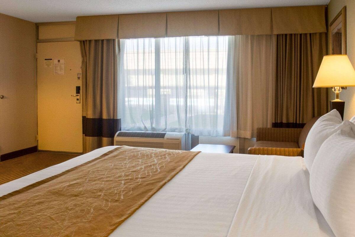 A hotel room with a large bed, a bedside table, a chair, a window with curtains, and an air conditioning unit.