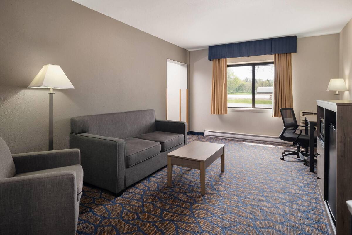 A hotel room with a sofa, armchair, desk, lamp, and a window with curtains.