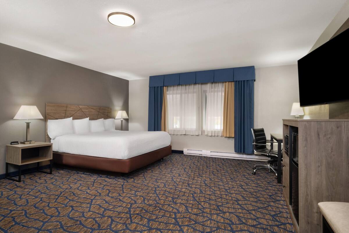 A hotel room with a large bed, two bedside tables with lamps, a flat-screen TV, a desk with a chair, and a patterned carpet.