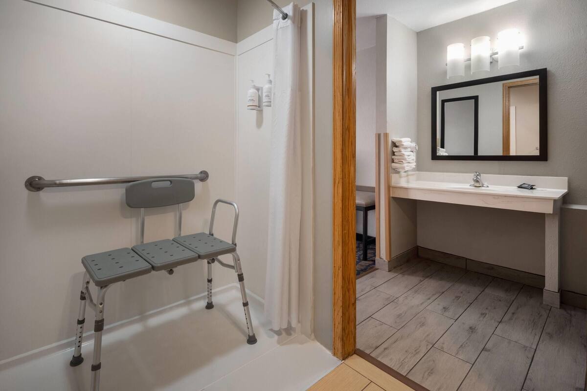 A bathroom with a shower, a handrail, a wheelchair, a sink, and a mirror.