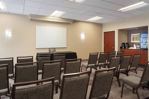 Una sala de conferencias con filas de sillas, una pizarra, una pantalla de proyector, una estación de café y un refrigerador.