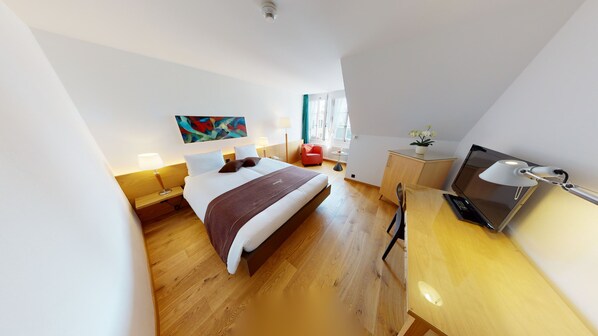 A modern hotel room with a large bed, a desk with a computer, a chair, a small table with a plant, and a colorful abstract painting on the wall.