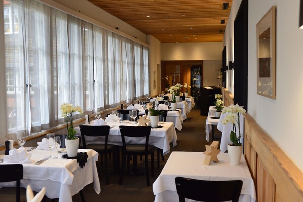 A restaurant with multiple tables set for dining, white tablecloths, and potted plants.