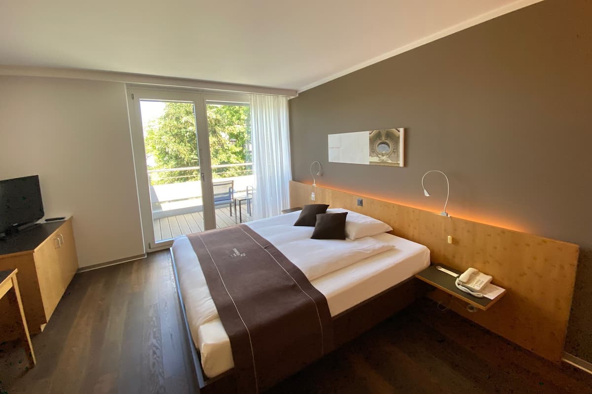 Habitación de hotel con cama, escritorio, silla, balcón con mesa y teléfono en la mesita de noche.