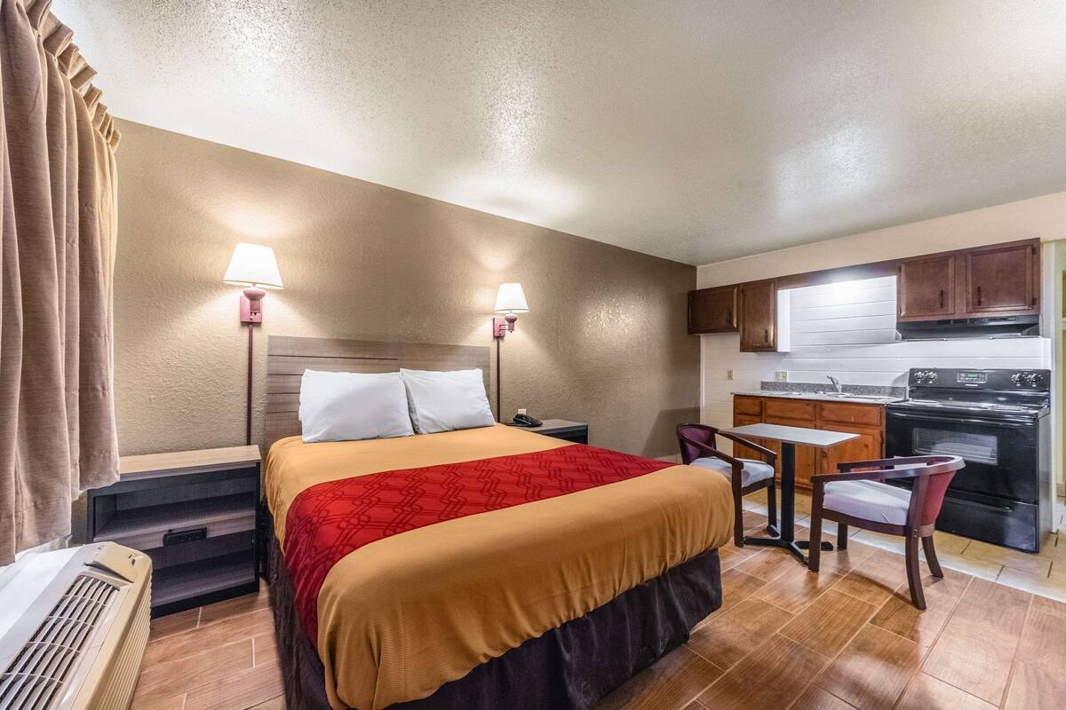 Habitación de hotel con cama, lámparas de noche, una pequeña mesa con sillas, kitchenette y ventana con cortinas.