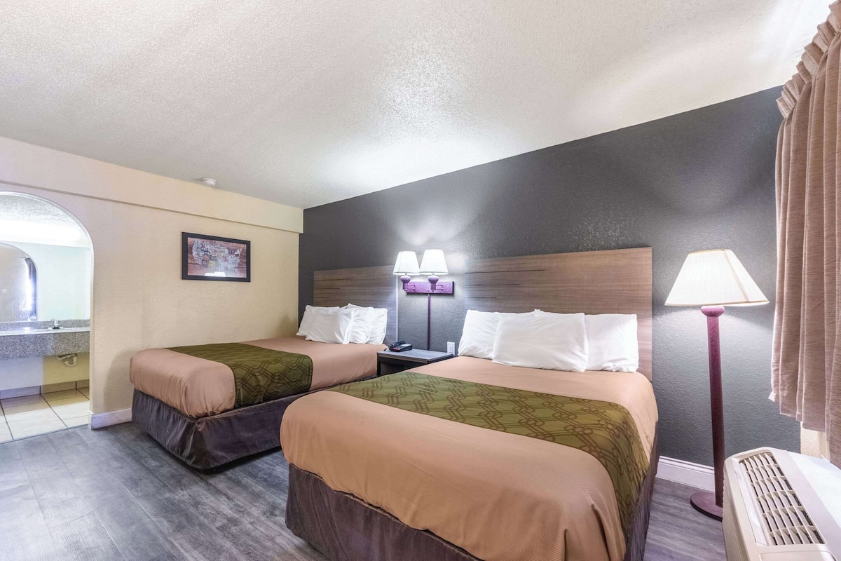 Habitación de hotel con dos camas, una mesita de noche, una lámpara, un cuadro en la pared y una ventana con cortinas.