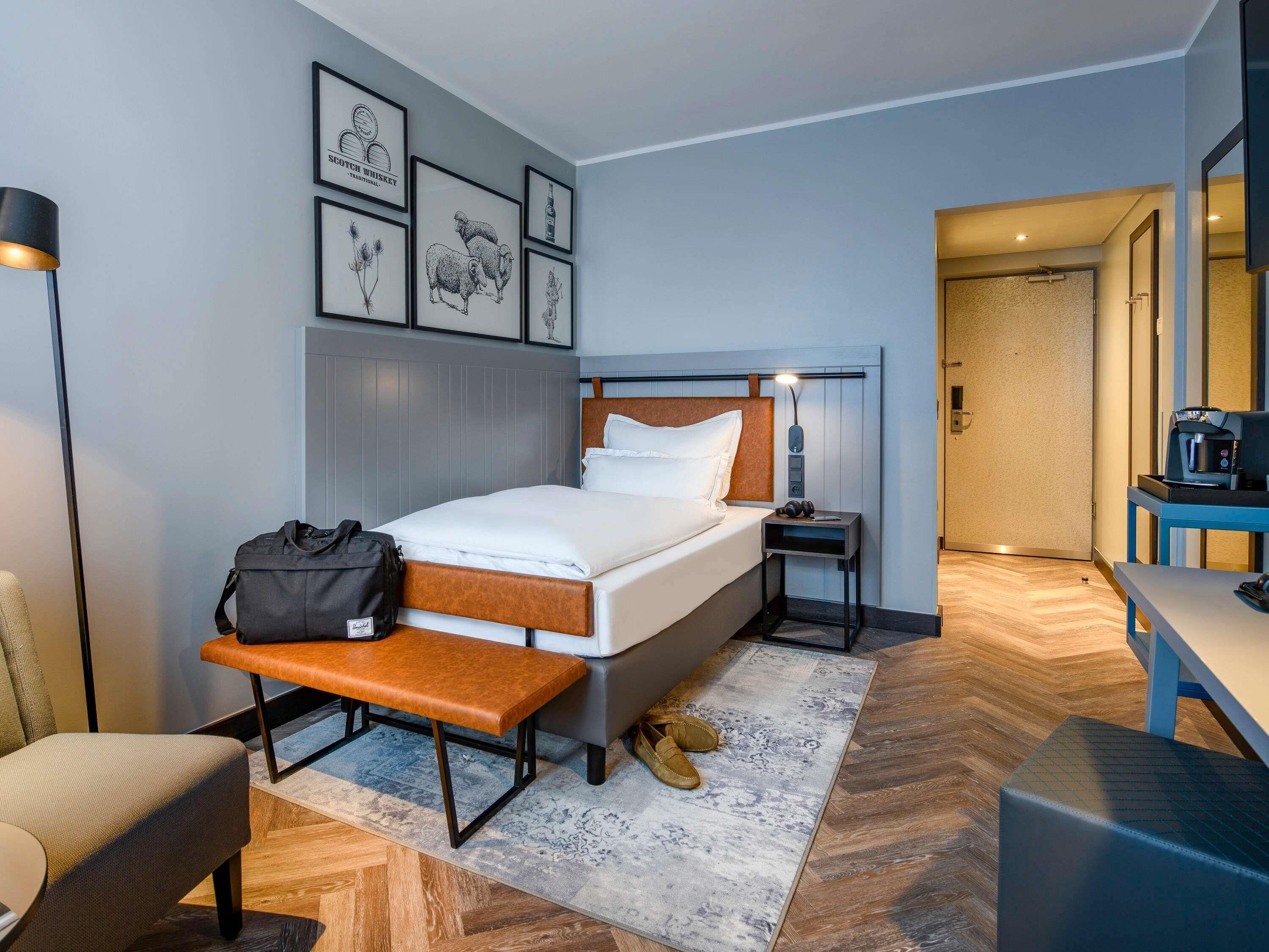 A hotel room with a bed, bedside table, lamp, bench, chair, and a desk with a coffee machine.