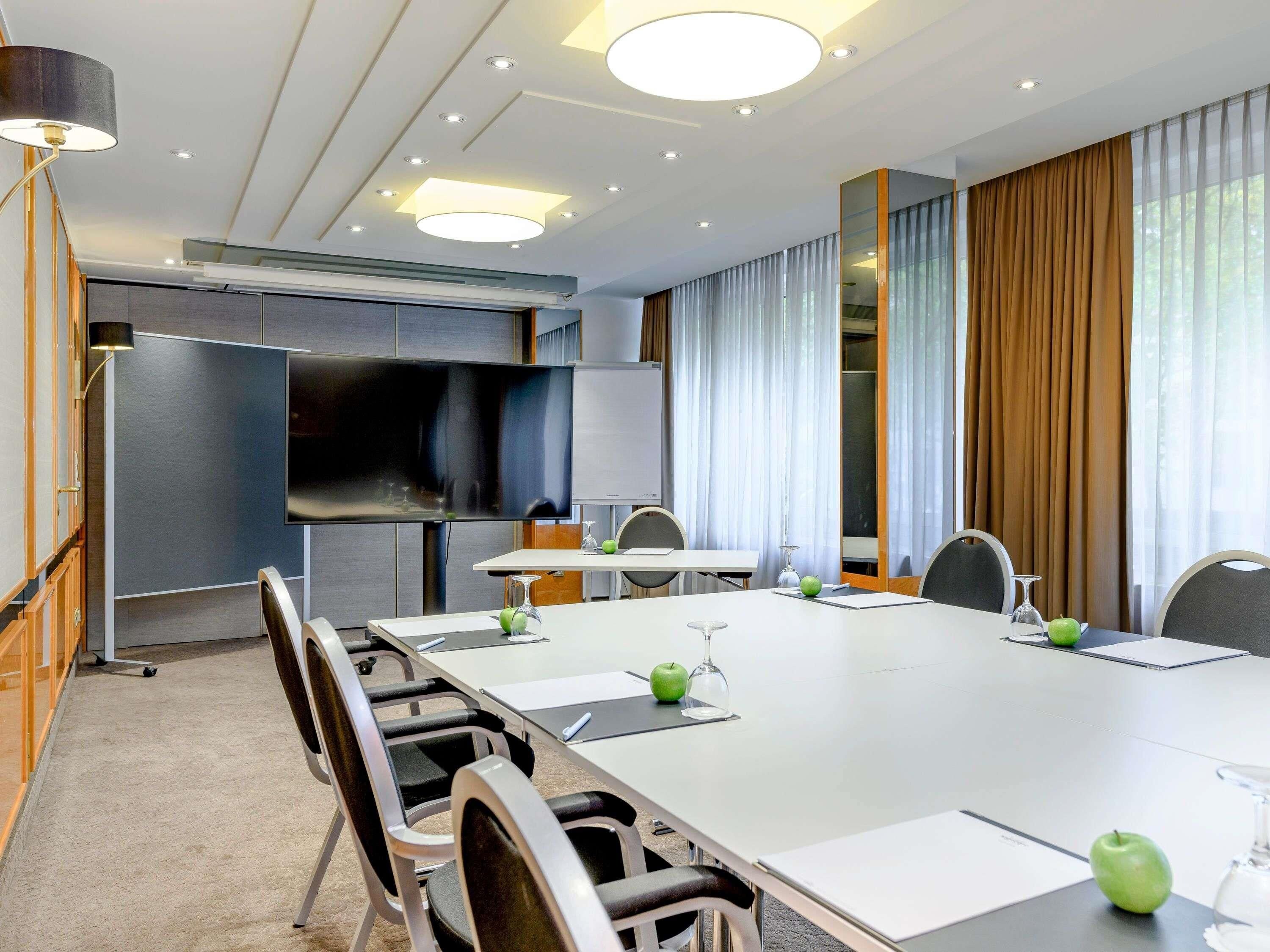 A conference room with a long table, chairs, a flat-screen TV, a whiteboard, and a large window with curtains.
