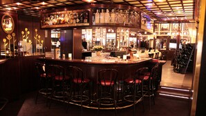 Zona de bar con mostrador curvo, varios taburetes y una variedad de botellas y vasos en estantes.