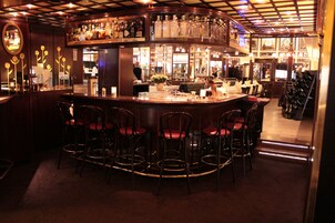 Zona de bar con mostrador curvo, varios taburetes y una variedad de botellas y vasos en estantes.